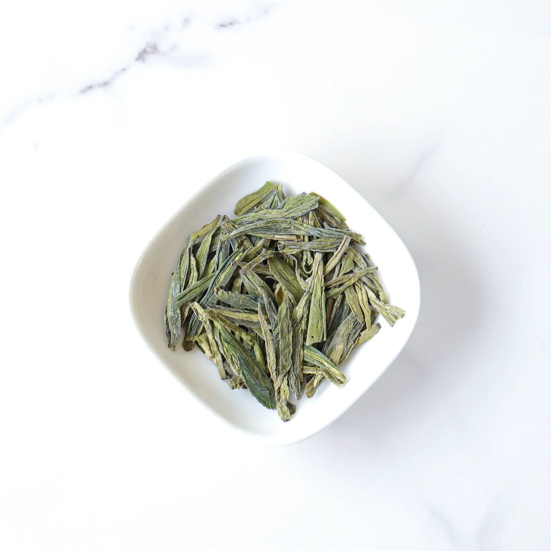 A white bowl of My Store’s Longjing Dragon Well Green Tea, celebrated for its delicate chestnut aroma, rests on a white marble surface.