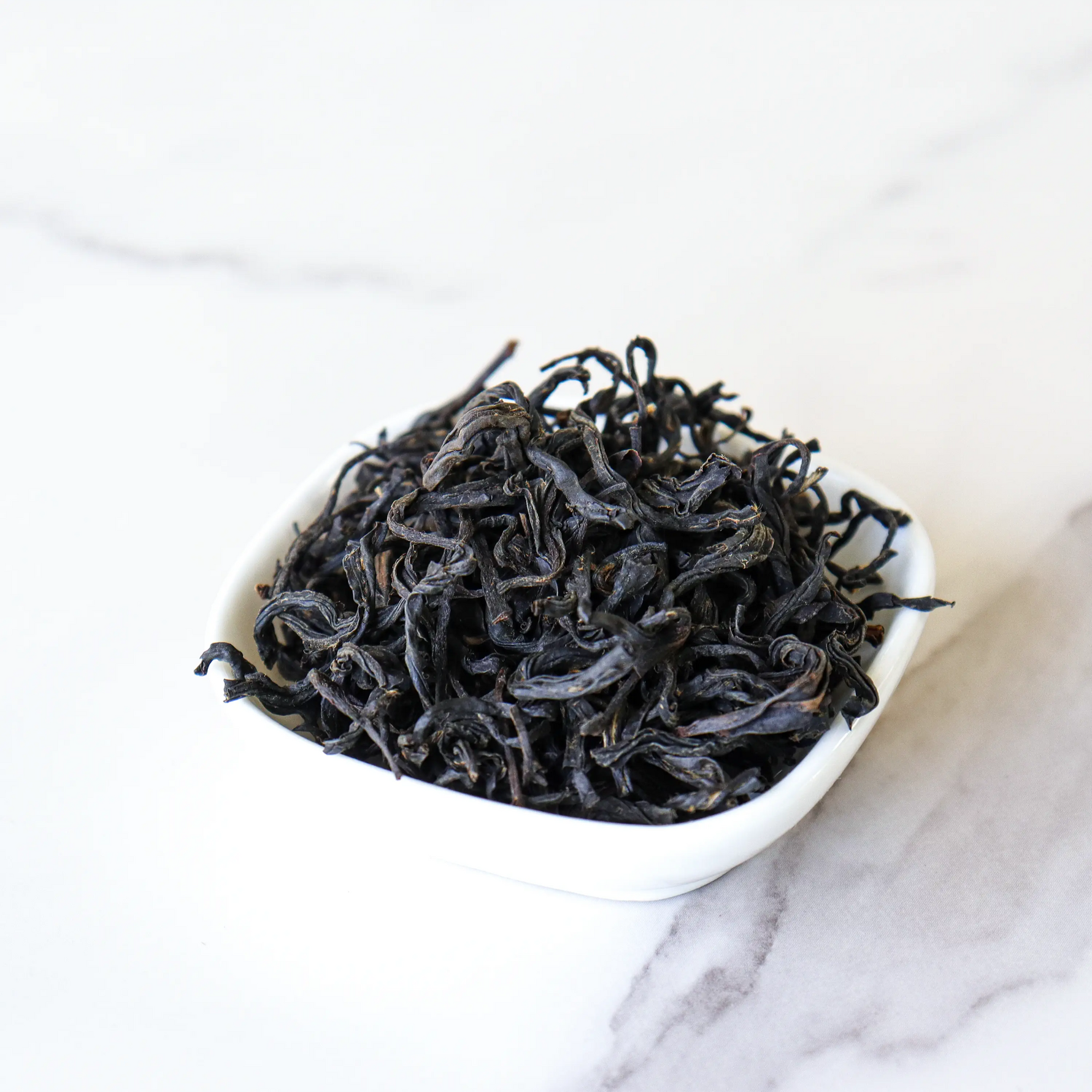 A small white bowl filled with loose, dried My Store's Oriental Beauty Oolong Tea leaves sits on a light-colored marble surface.
