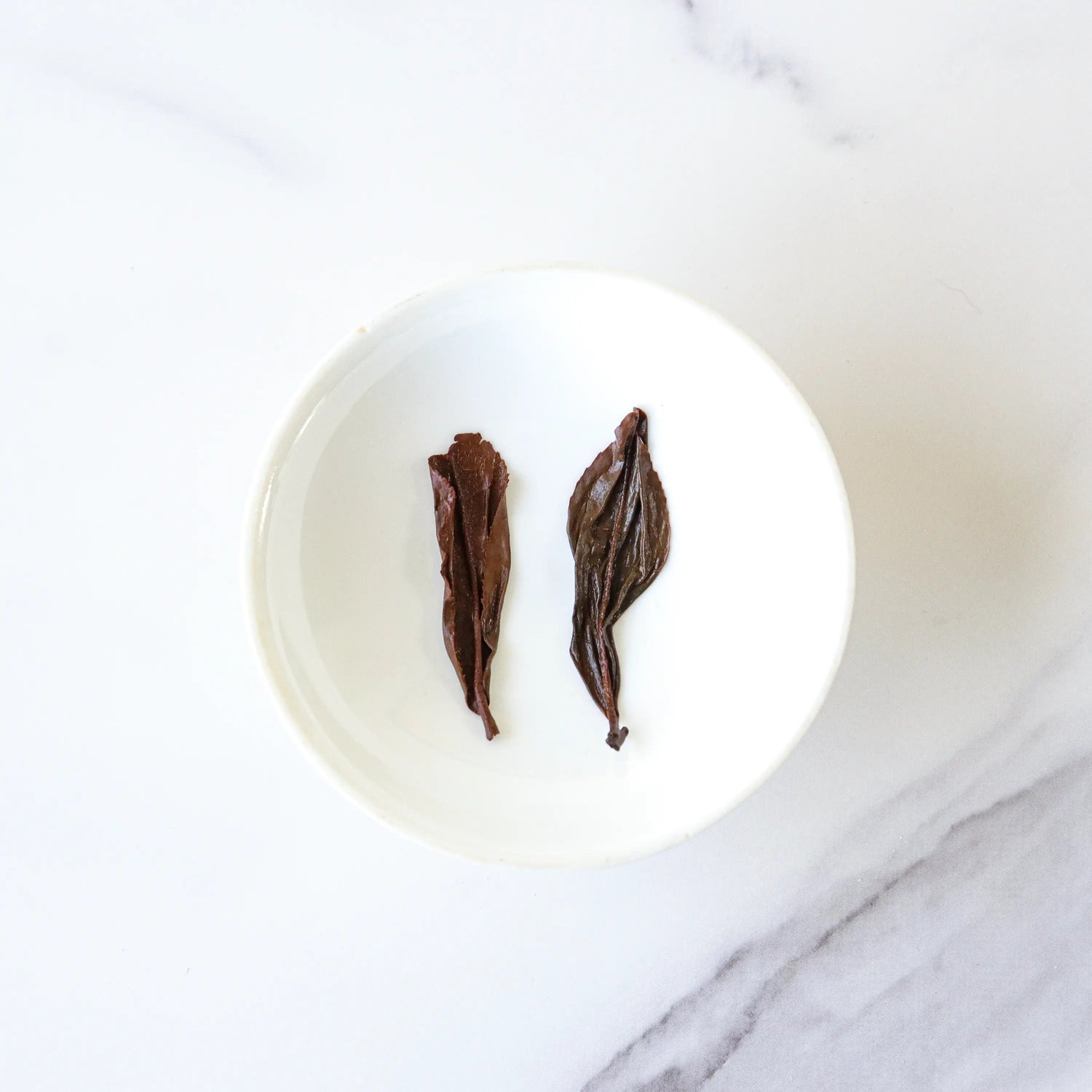 Two dried Oriental Beauty Oolong Tea leaves from My Store are placed side by side in a small white bowl on a white marble surface.