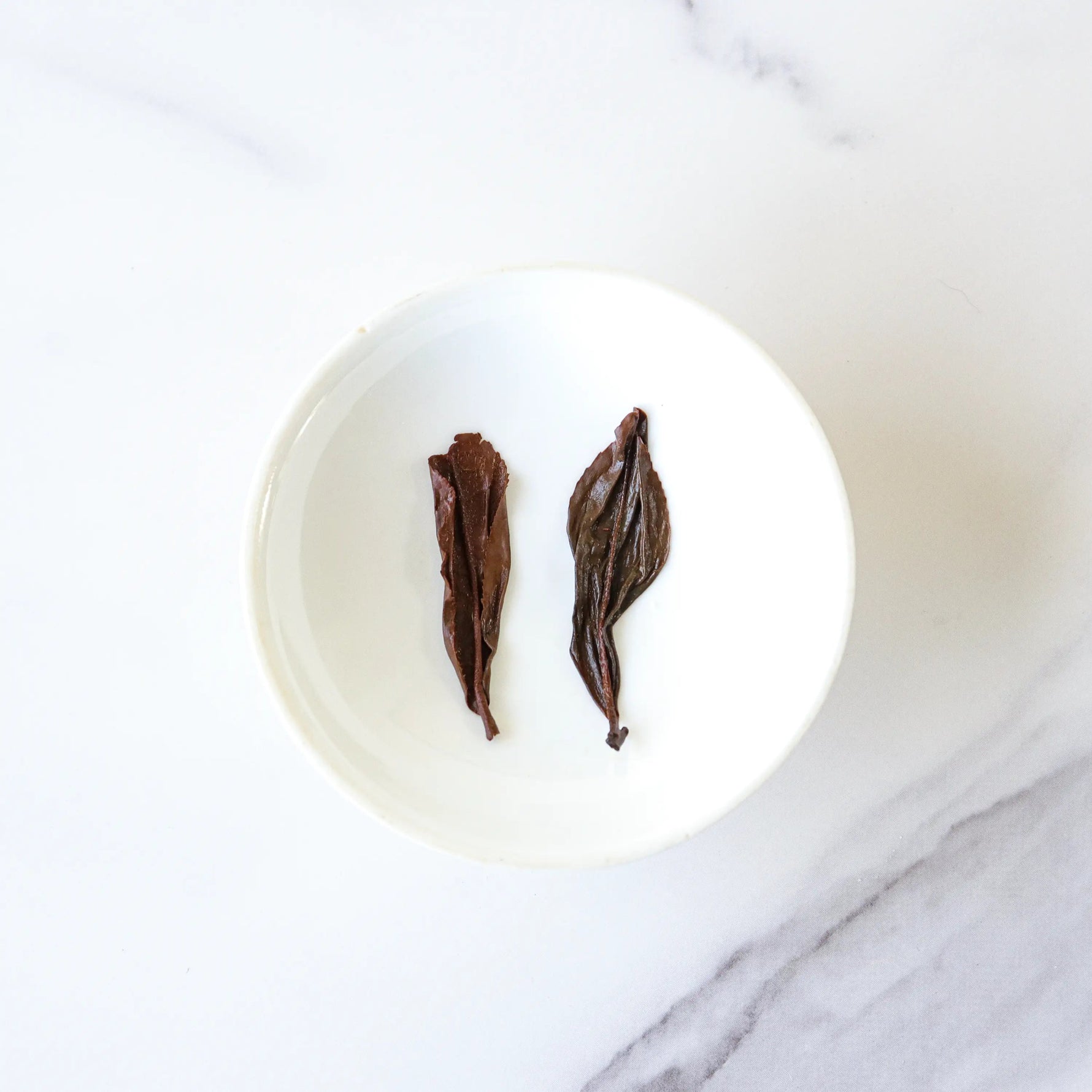Two dried Oriental Beauty Oolong Tea leaves from My Store are placed side by side in a small white bowl on a white marble surface.