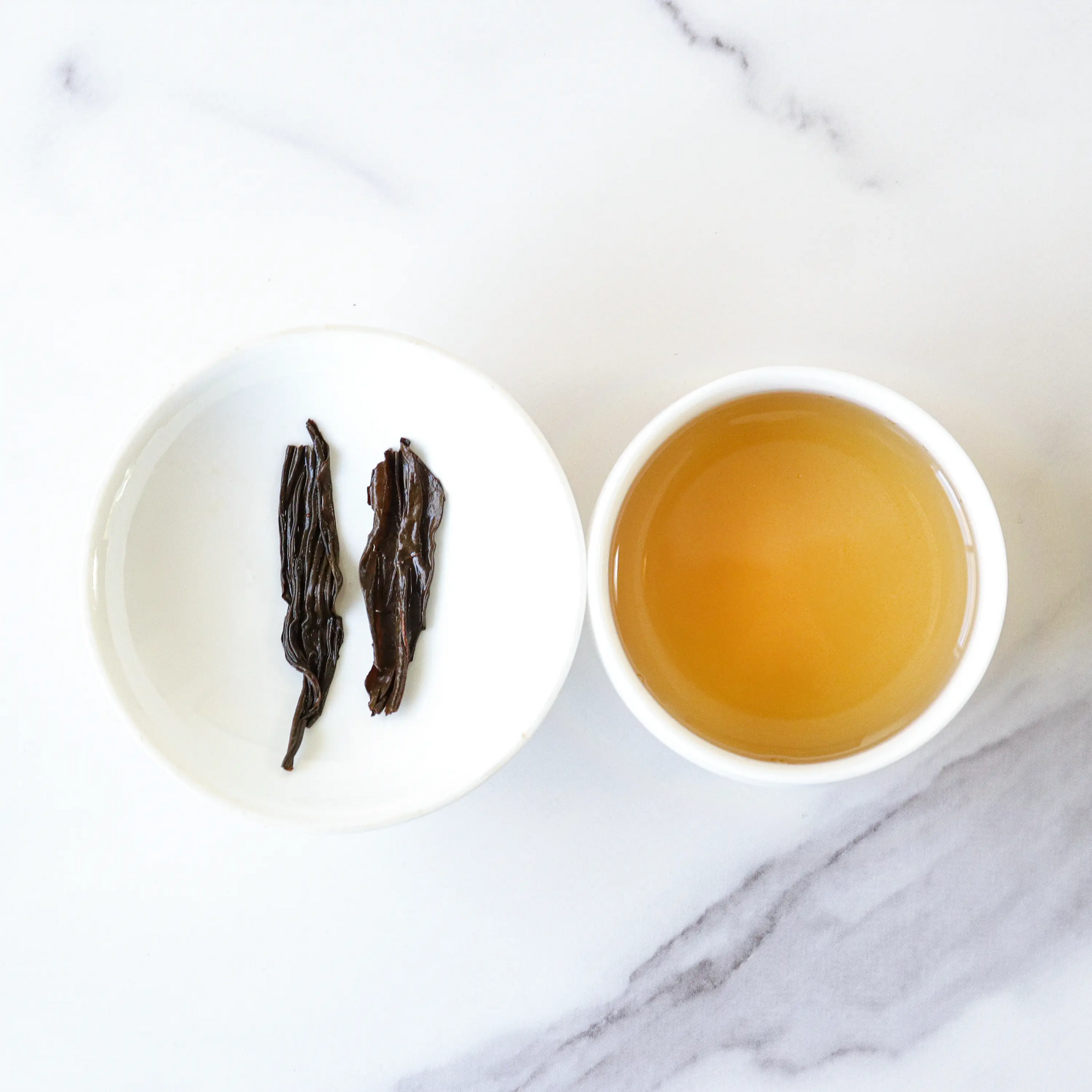 A small white dish with two Phoenix Dancong Oolong Tea leaves from My Store sits beside a cup of light amber tea on a white marble surface, showcasing the tea’s delicate floral fragrance.