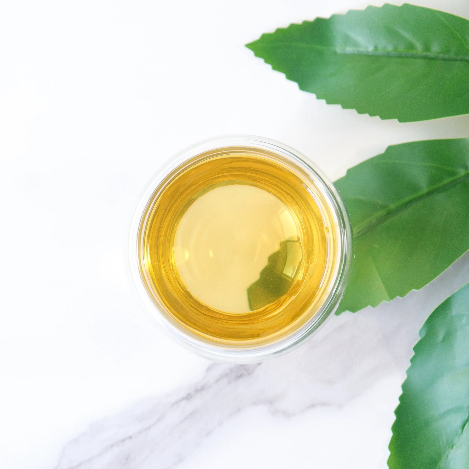 A glass of Pomelo Flower Green Tea from My Store rests on a white surface with green leaves nearby, evoking a serene nod to wellness tea traditions.