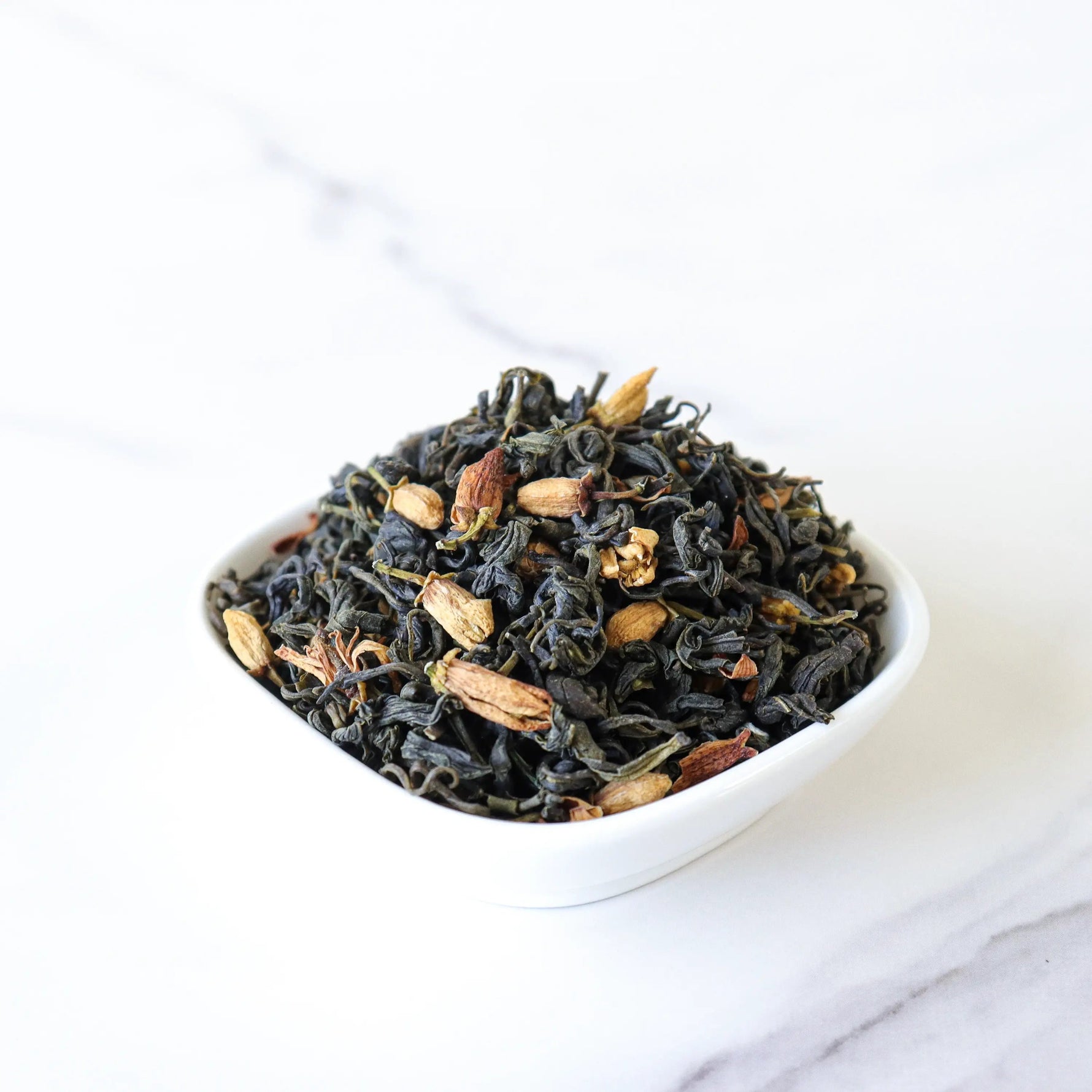 A small white bowl filled with Pomelo Flower Green Tea by My Store, a blend of loose leaf green tea and dried spices, rests on a white marble surface—ideal for anyone seeking a soothing wellness tea experience.