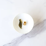 A white bowl holding a green leaf, a brown nut, and a light brown piece sits on white marble—perfect for savoring My Store's Pomelo Flower Green Tea.