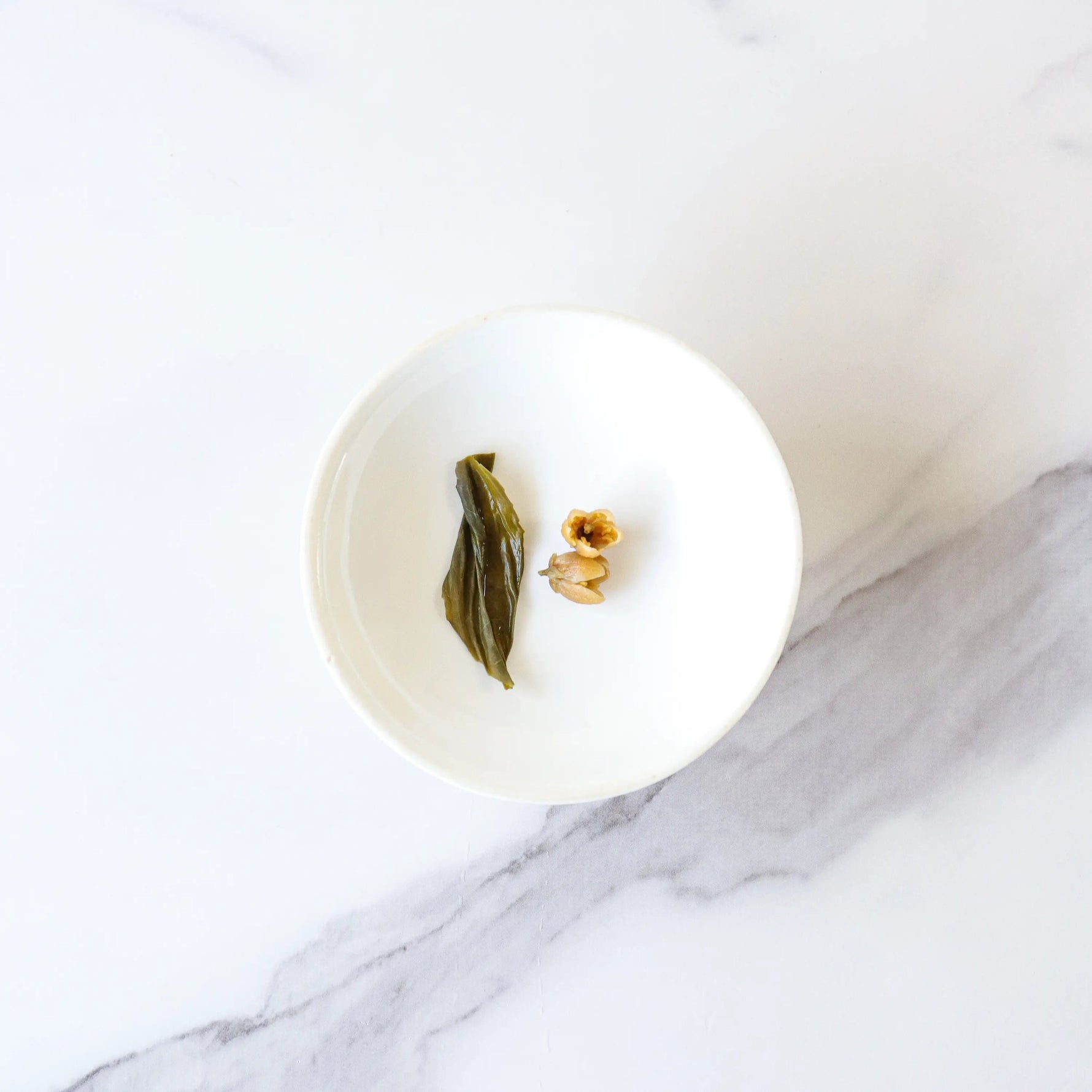 A white bowl holding a green leaf, a brown nut, and a light brown piece sits on white marble—perfect for savoring My Store's Pomelo Flower Green Tea.
