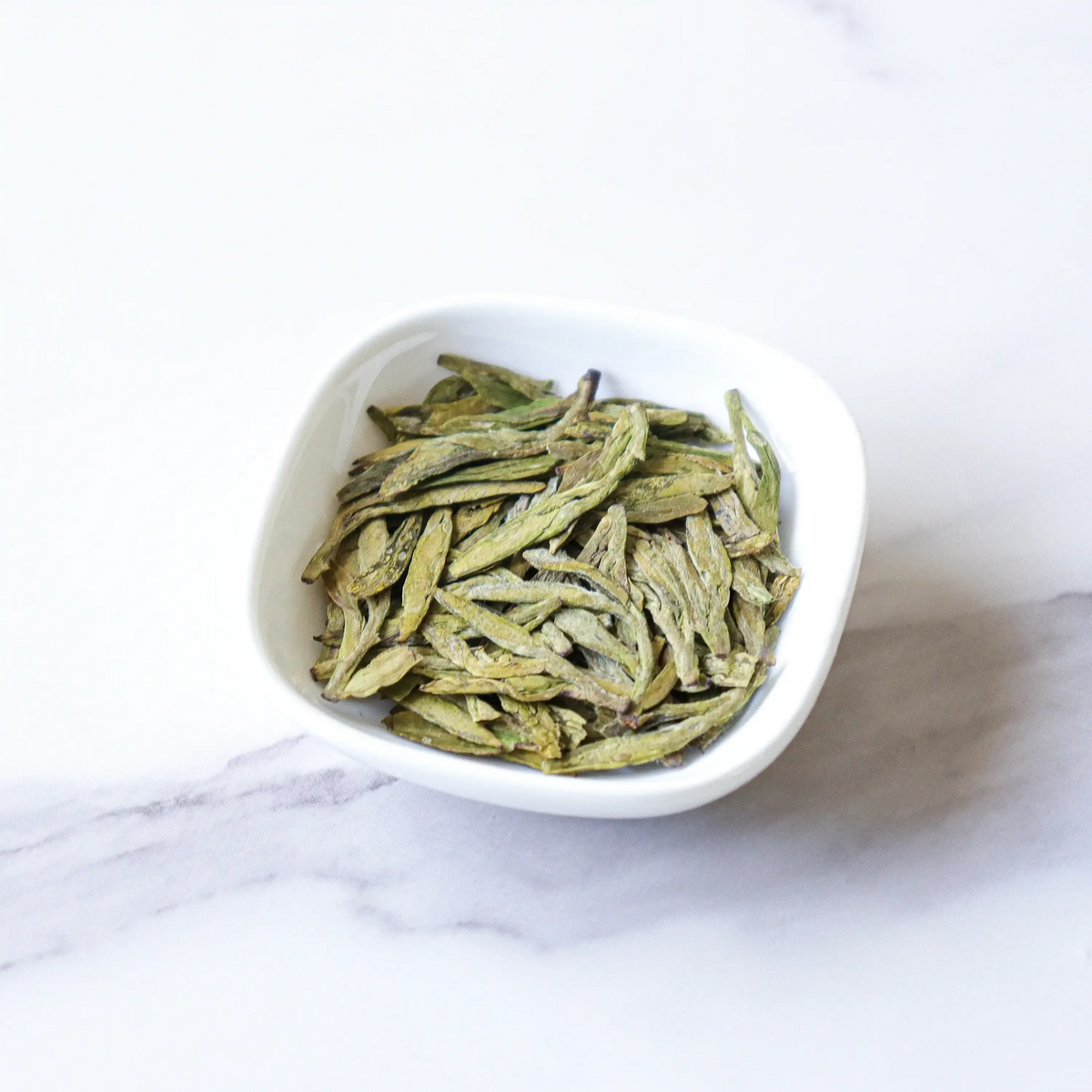Premium West Lake Longjing Tea flat green leaves in a white bowl on marble.