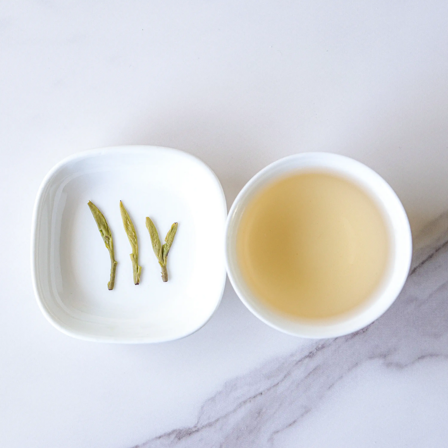 A white dish with three My Store Xihu Longjing Dragon Well Green Tea leaves sits next to a cup of light-colored tea, both resting on a white marble surface.