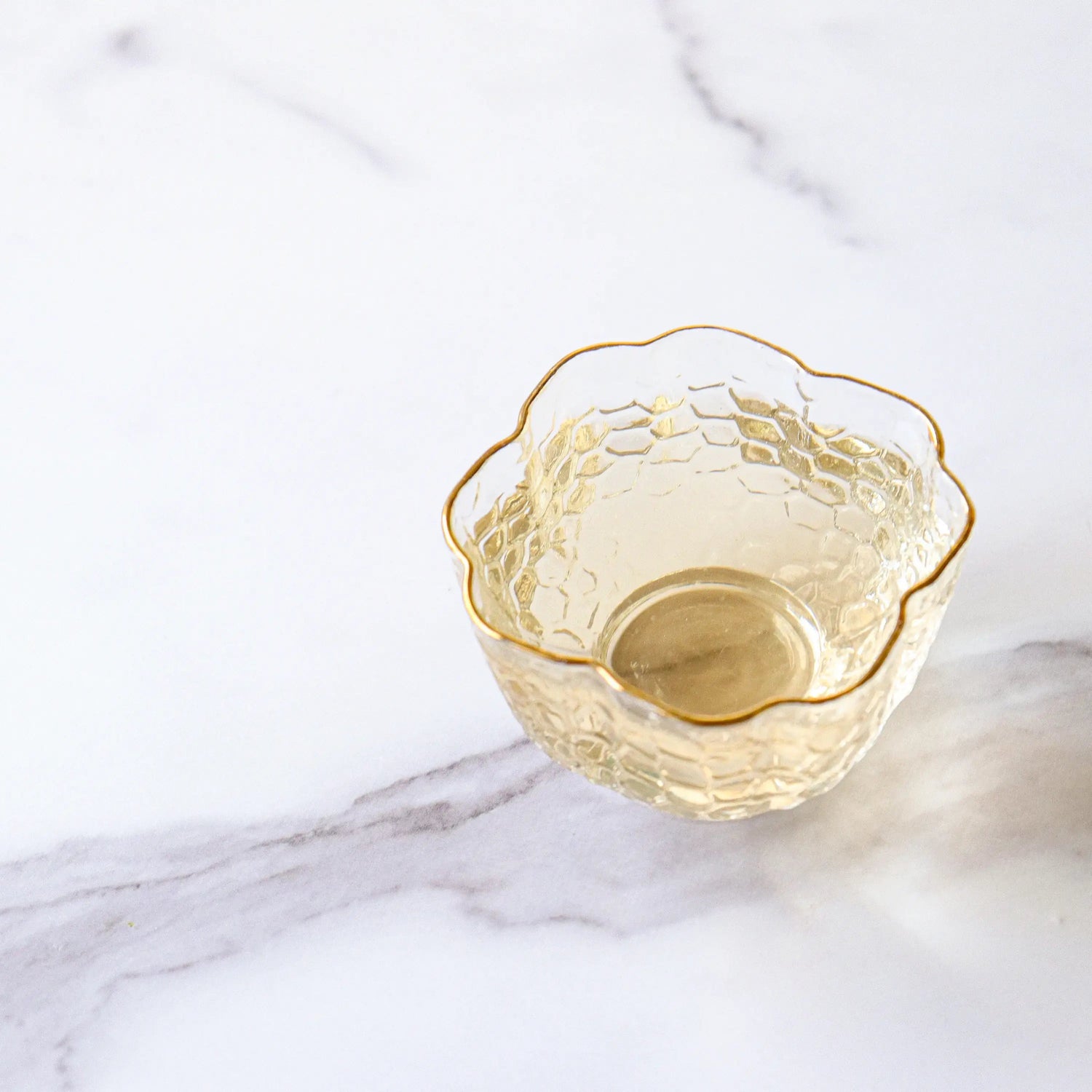 A small, empty, translucent glass bowl with a honeycomb pattern and scalloped rim sits on white marble, perfect for serving My Store's Xihu Longjing Dragon Well Green Tea or your favorite wellness tea.