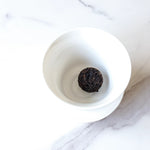 A Ripened Dragon Pu'er Tea Pearl by My Store from Yunnan Province rests in a white porcelain bowl on a marble surface.