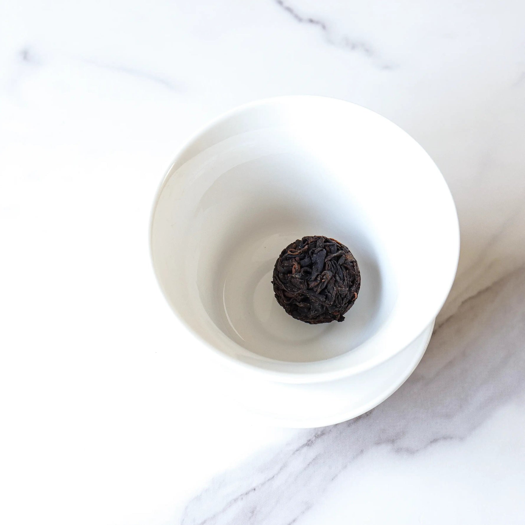 A Ripened Dragon Pu'er Tea Pearl by My Store from Yunnan Province rests in a white porcelain bowl on a marble surface.
