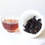 A glass cup of dark Ripened Dragon Pu'er Tea Pearls from My Store sits beside a white bowl of loose, steeped leaves, all on a white background.