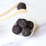 A small white dish displays five dark, round Ripened Dragon Pu'er Tea Pearls from My Store. Wooden tongs lift one pearl above the dish, set against a light marble surface inspired by Yunnan tea traditions.