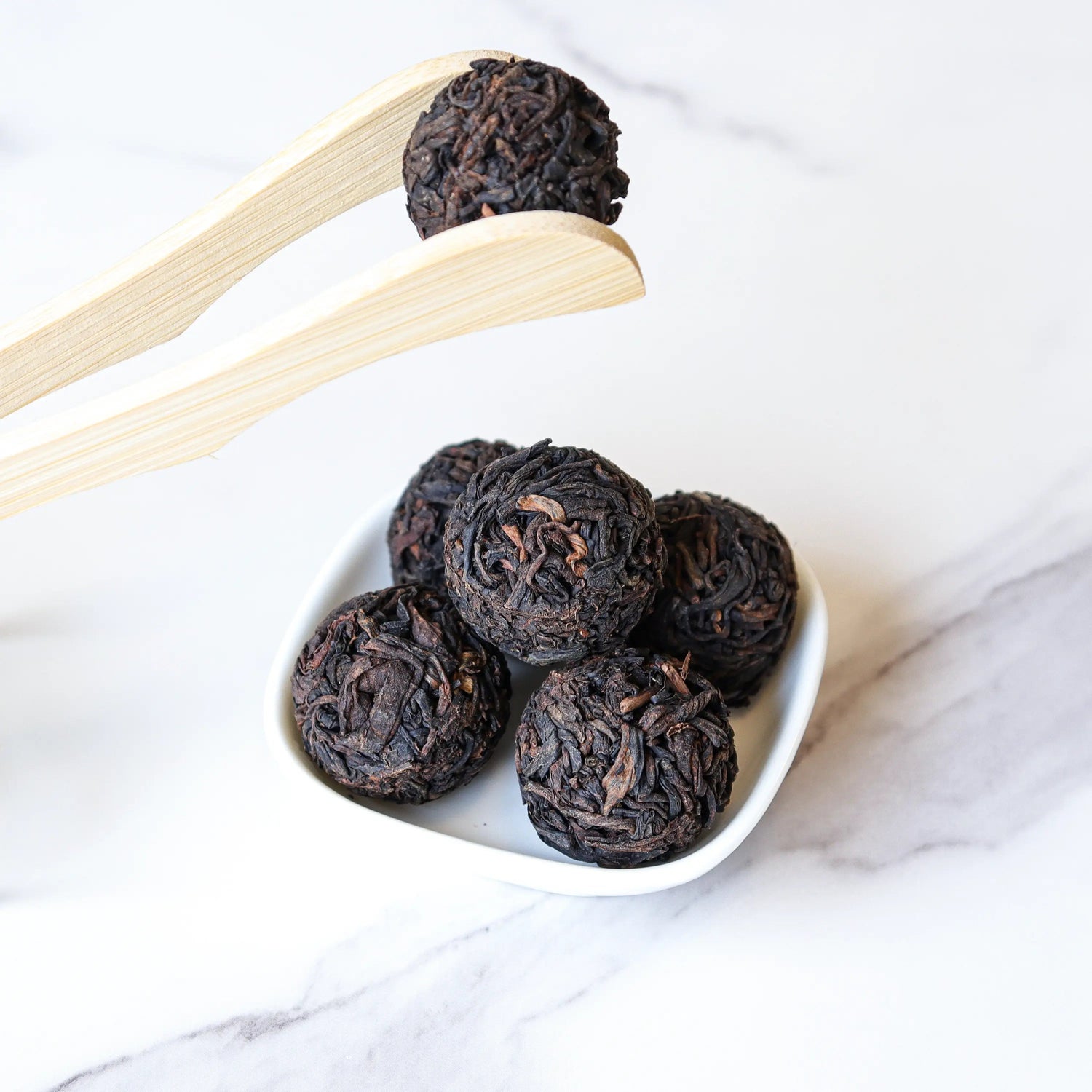 A small white dish displays five dark, round Ripened Dragon Pu'er Tea Pearls from My Store. Wooden tongs lift one pearl above the dish, set against a light marble surface inspired by Yunnan tea traditions.