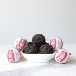 A small white bowl holds six dark Ripened Dragon Pu'er Tea Pearls by My Store, with six individually wrapped tea balls in white and red paper, all displayed on a white surface—an elegant taste of Yunnan Province.