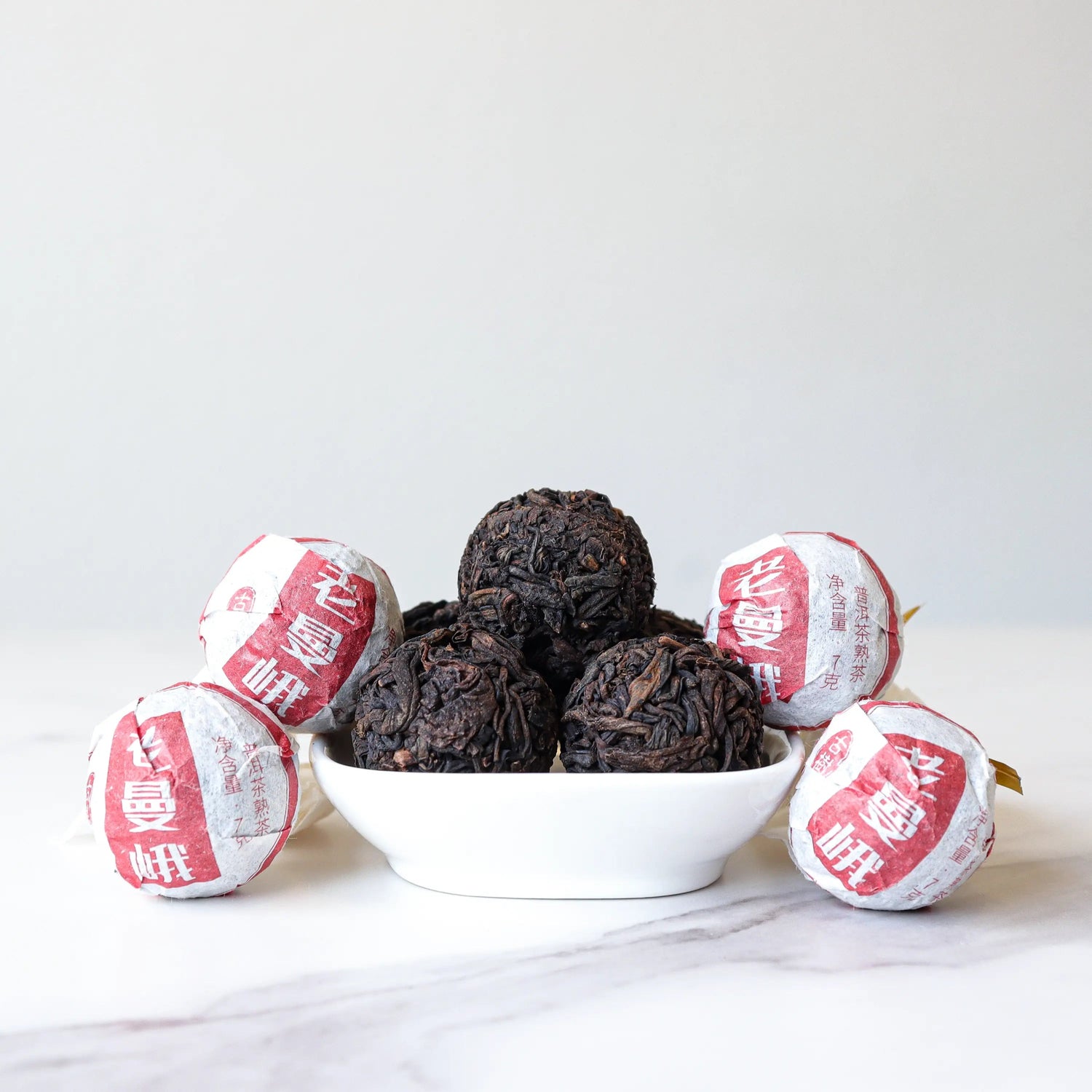 A small white bowl holds six dark Ripened Dragon Pu'er Tea Pearls by My Store, with six individually wrapped tea balls in white and red paper, all displayed on a white surface—an elegant taste of Yunnan Province.