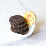 A small white dish holds four stacked My Store Small Ripened Pu'er Tea Cakes; a wrapped premium tea cake with Chinese text stands upright behind them on a white marble surface, showcasing their Yunnan Province origin.