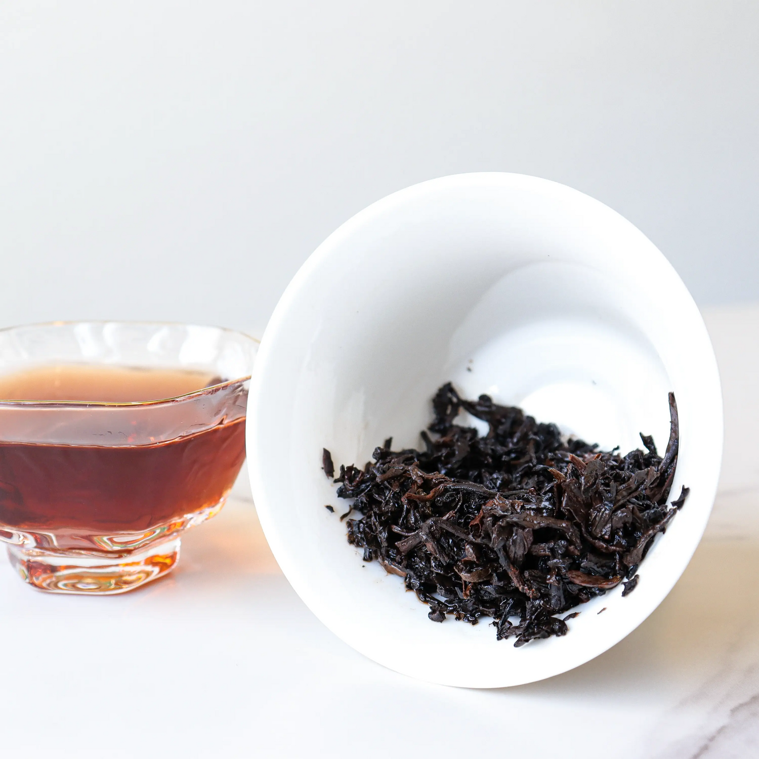 A glass cup of brewed tea sits next to a white bowl holding loose Small Ripened Pu'er Tea Cakes by My Store, all arranged on a pristine white surface.