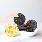 A small stack of My Store Small Ripened Pu'er Tea Cakes from Yunnan rests in a white bowl, one held by a wooden utensil, with a yellow-labeled My Store tea tin beside them on a white surface.
