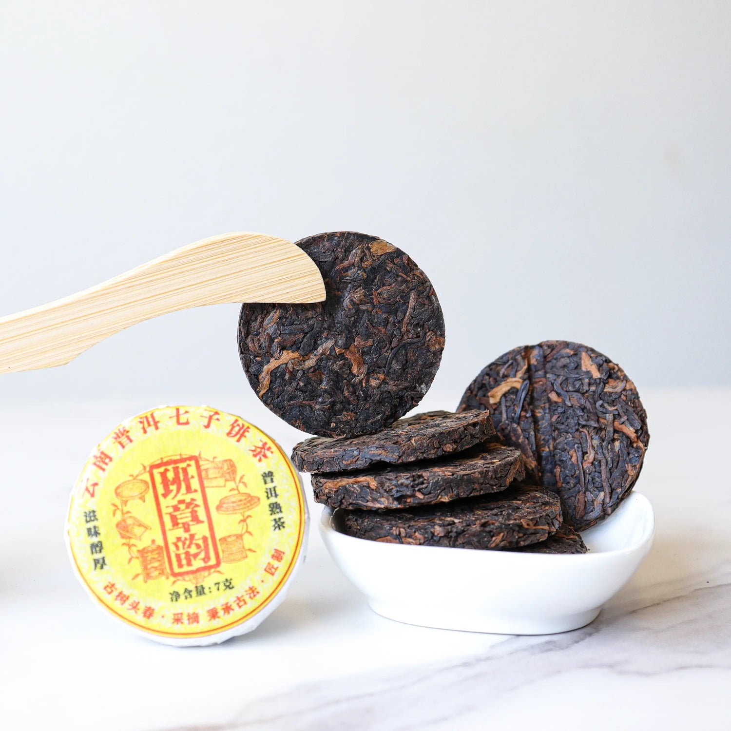 A small stack of My Store Small Ripened Pu'er Tea Cakes from Yunnan rests in a white bowl, one held by a wooden utensil, with a yellow-labeled My Store tea tin beside them on a white surface.