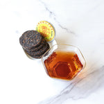A glass cup of amber-hued Small Ripened Pu'er Tea Cakes by My Store sits beside a dish of round tea cakes with yellow and red labels on white marble, showcasing this premium Chinese tea from Yunnan Province.