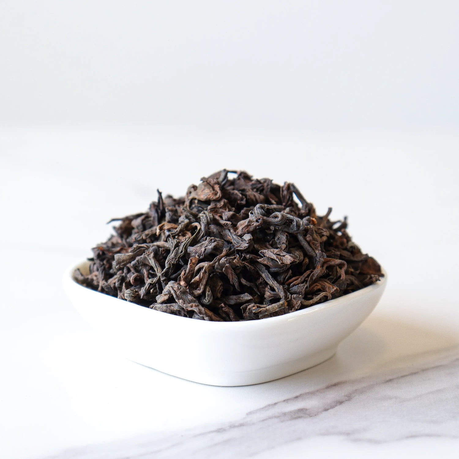 A white bowl of My Store Ripened Pu'er Tea sits on a marble surface against a plain white background, showcasing its unique fermentation process and origin from Yunnan Province.