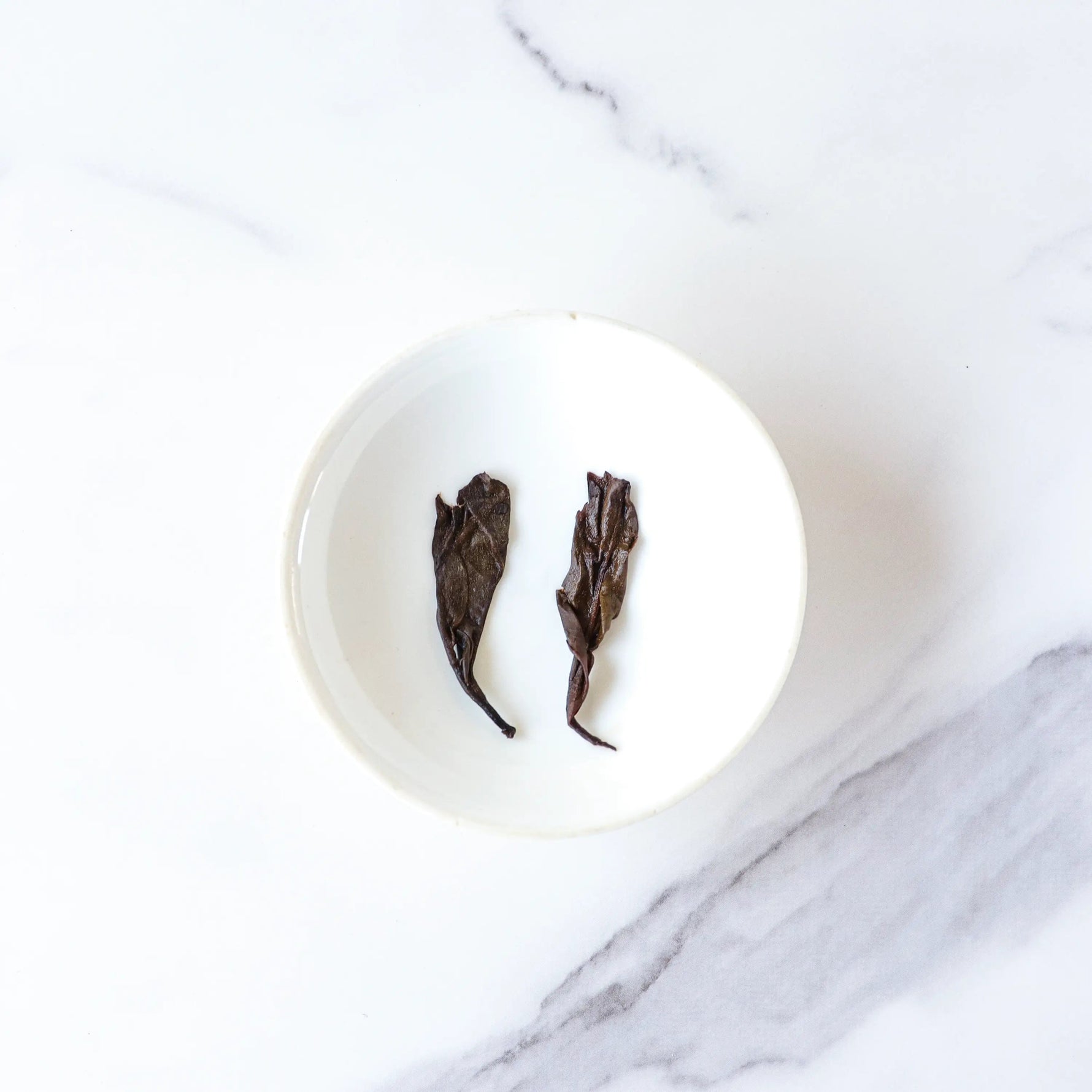 Two Ripened Pu'er Tea leaves from My Store rest side by side in a small white bowl on a white marble surface.
