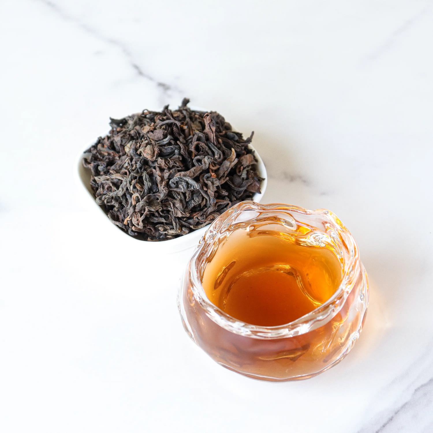A clear glass cup of amber-colored Ripened Pu'er Tea from My Store sits beside a white bowl of loose tea leaves, showcasing the rich traditions and unique fermentation process of Yunnan Province on a white surface.