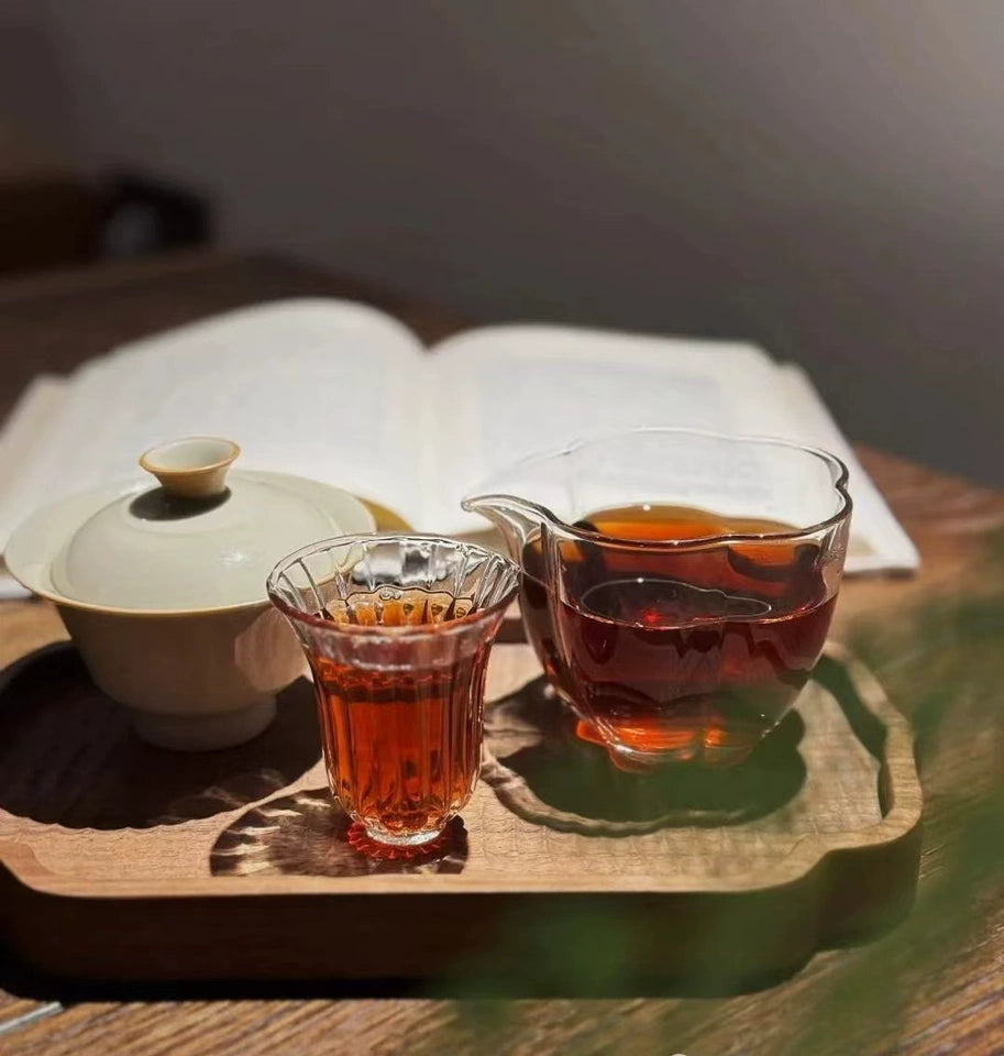 A wooden tray holds a glass pitcher and a small glass of tea beside a lidded teacup, with an open book in the blurred background.
