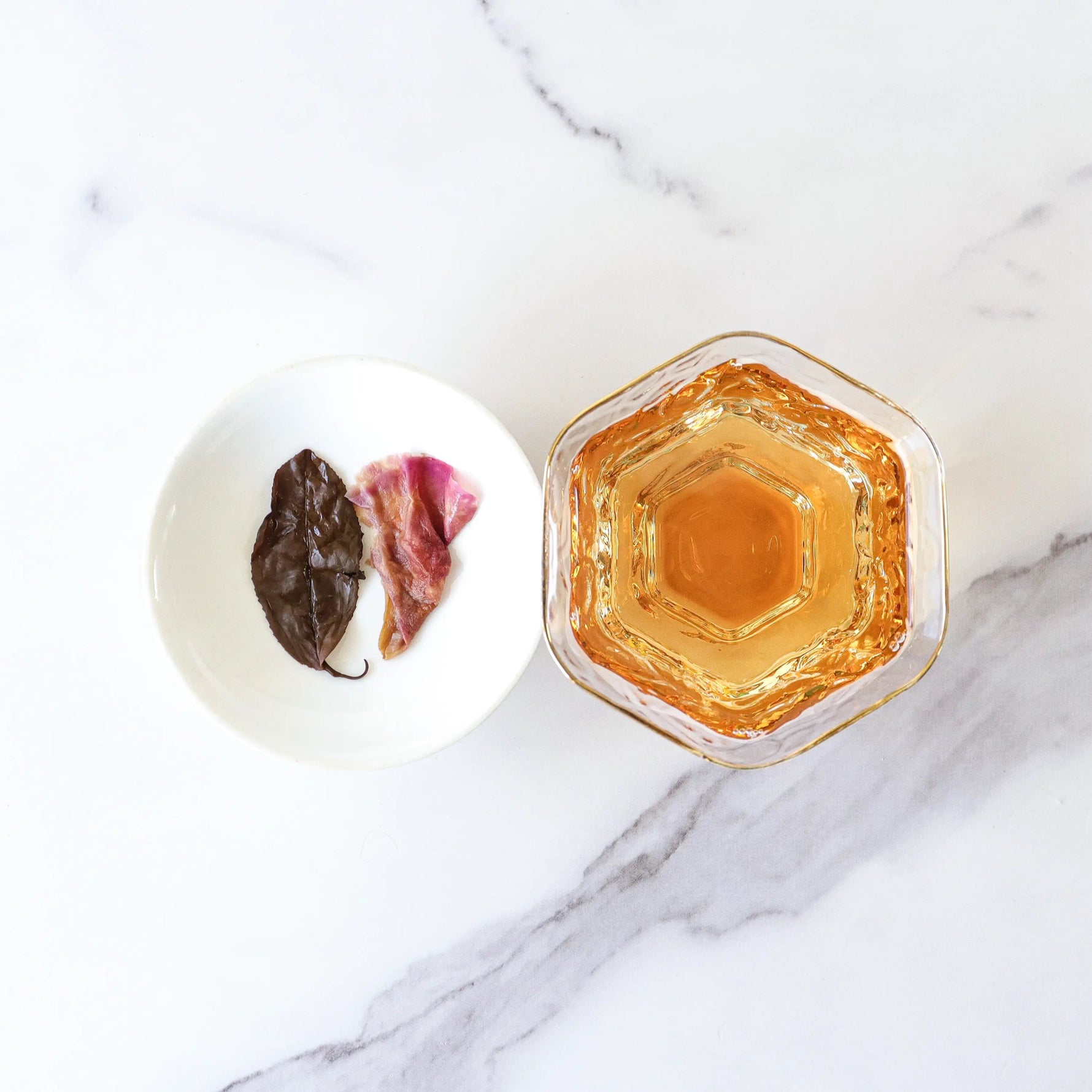 A glass of amber-colored Dianhong Rose Black Tea from My Store sits beside a small white dish holding two wet tea leaves on a white marble surface.