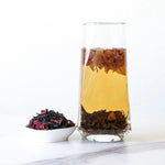 A glass of brewed My Store Dianhong Rose Black Tea, with tea leaves floating inside, sits beside a small white dish holding loose leaf tea and dried rose petals from Yunnan Province on a white surface.