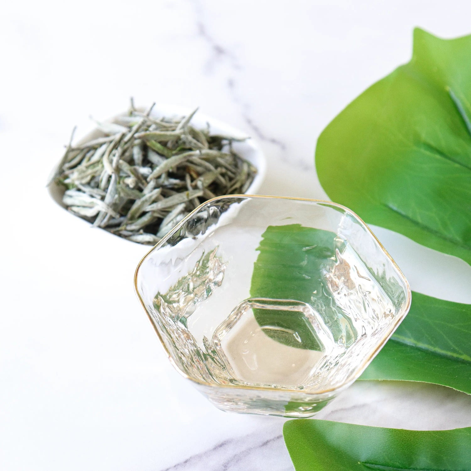 A glass cup rests on marble beside a white bowl of My Store's Silver Needle White Tea and large green leaves, showcasing the delicate beauty and antioxidants of this premium white tea.