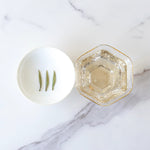 A white bowl with three tea leaves sits beside a hexagonal glass filled with My Store Silver Needle White Tea, known for its antioxidants, on a marble surface.