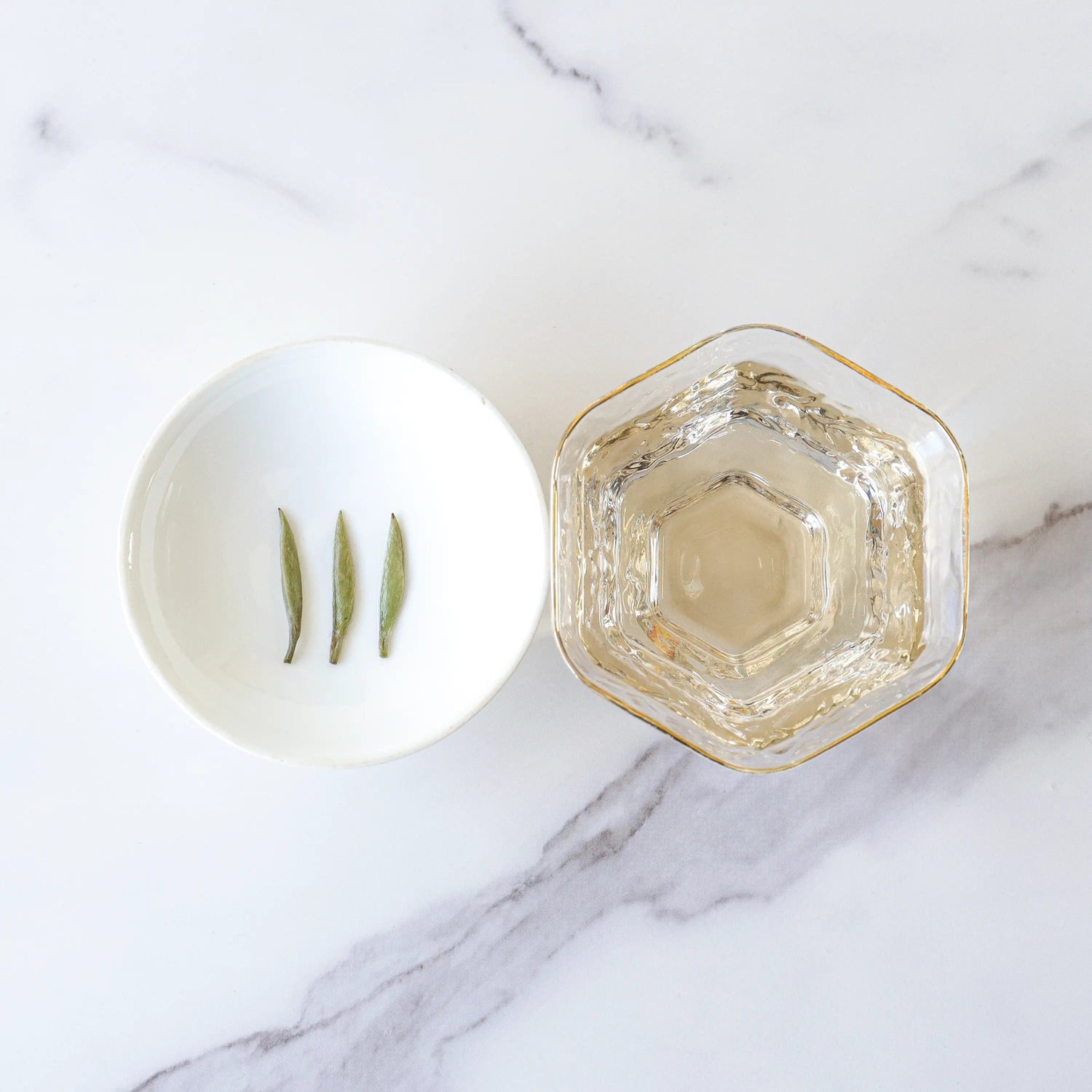 A white bowl with three tea leaves sits beside a hexagonal glass filled with My Store Silver Needle White Tea, known for its antioxidants, on a marble surface.