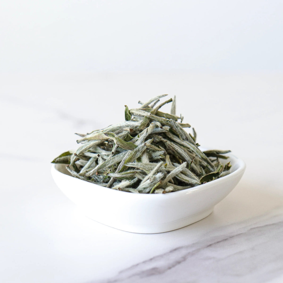 Silver Needle White Tea buds in a white bowl on a clean white surface.