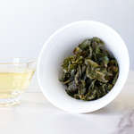 A white bowl holds steeped My Store Tie Guan Yin Oolong Tea leaves beside a glass cup of light yellow tea, prized for its delicate orchid aroma, on a white surface.
