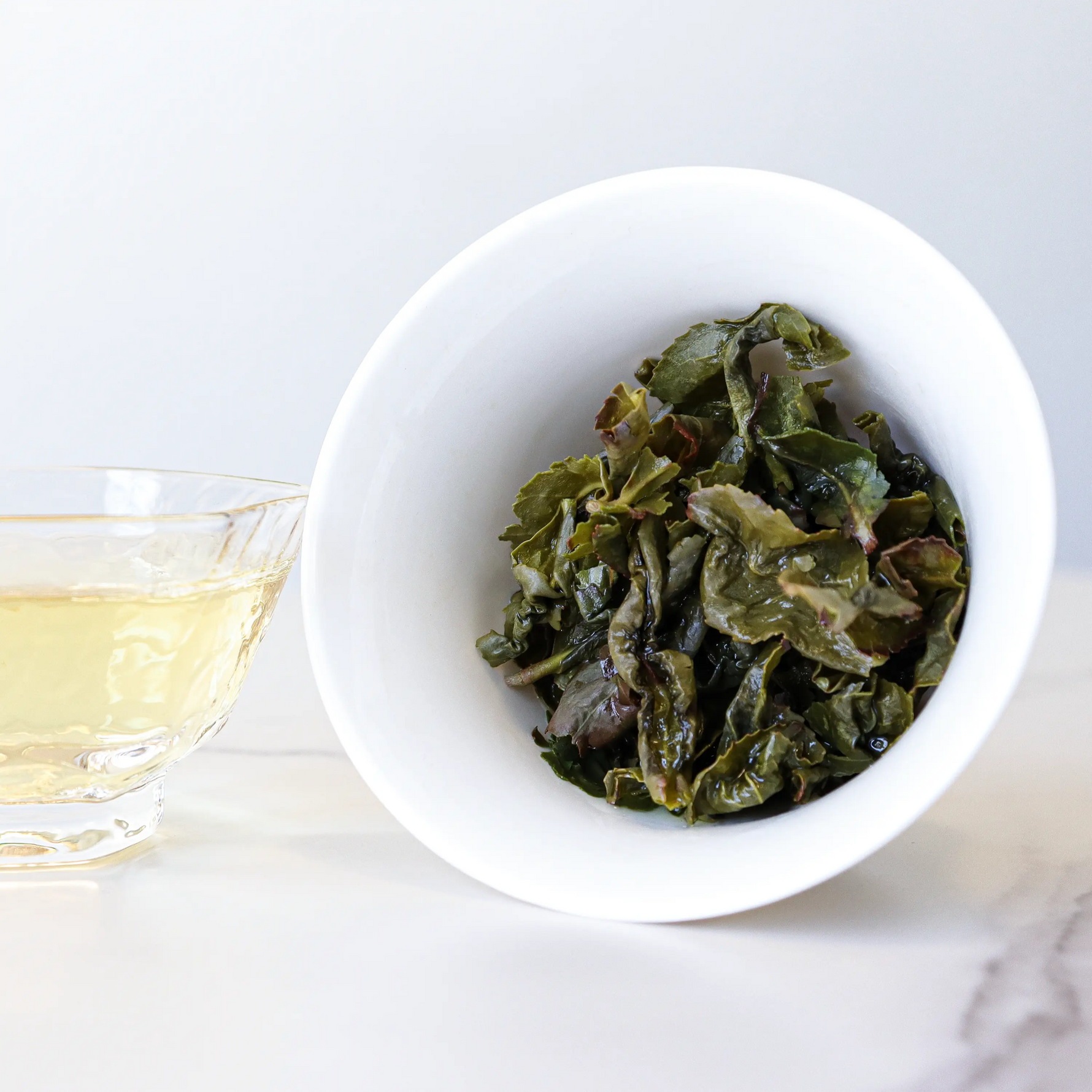 A white bowl holds steeped My Store Tie Guan Yin Oolong Tea leaves beside a glass cup of light yellow tea, prized for its delicate orchid aroma, on a white surface.