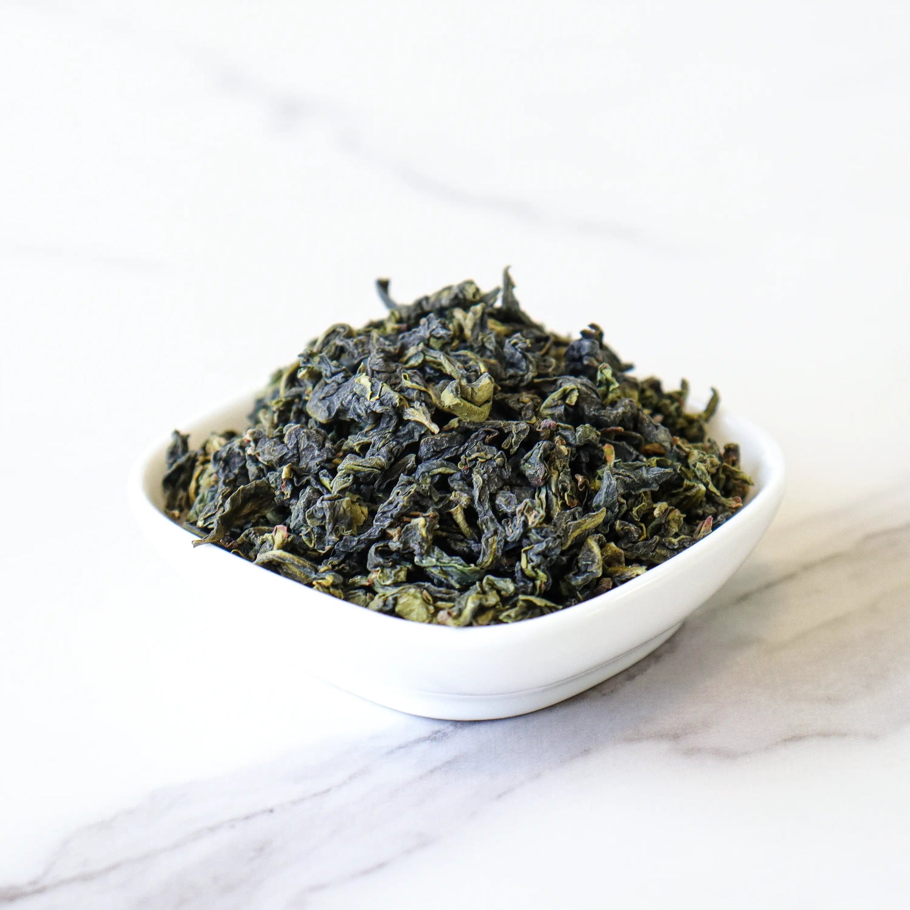 A small white bowl filled with My Store's Tie Guan Yin Oolong Tea, known for its subtle orchid aroma, rests on a white marble surface.