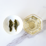 A glass cup of My Store's Tie Guan Yin Oolong Tea next to a white dish with two steeped tea leaves, arranged on a white marble surface.