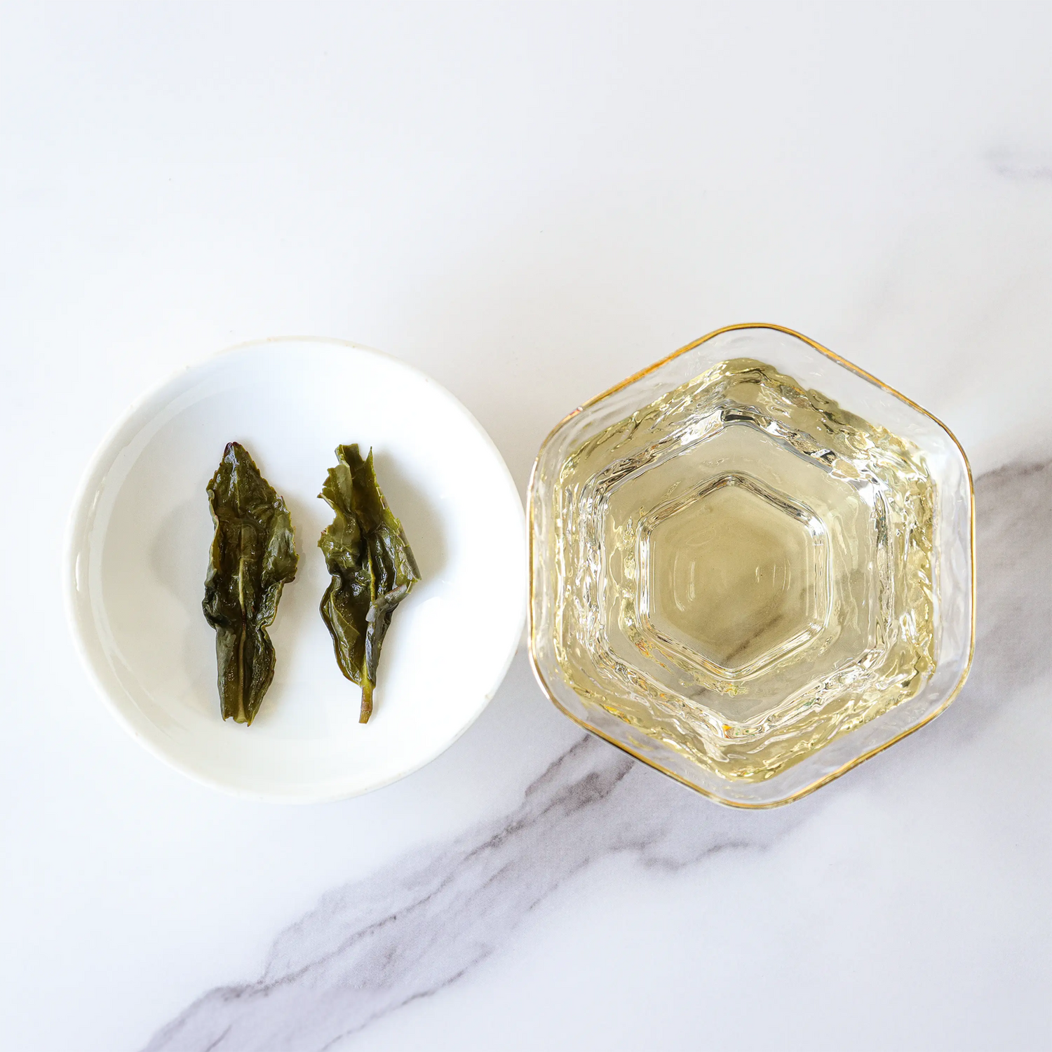 A glass cup of My Store's Tie Guan Yin Oolong Tea next to a white dish with two steeped tea leaves, arranged on a white marble surface.