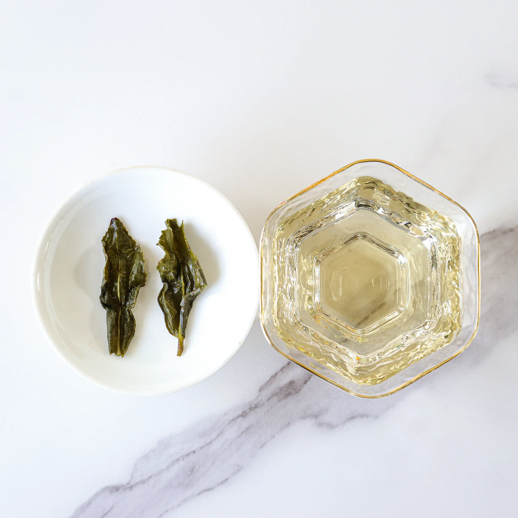 A glass cup of My Store's Tie Guan Yin Oolong Tea next to a white dish with two steeped tea leaves, arranged on a white marble surface.