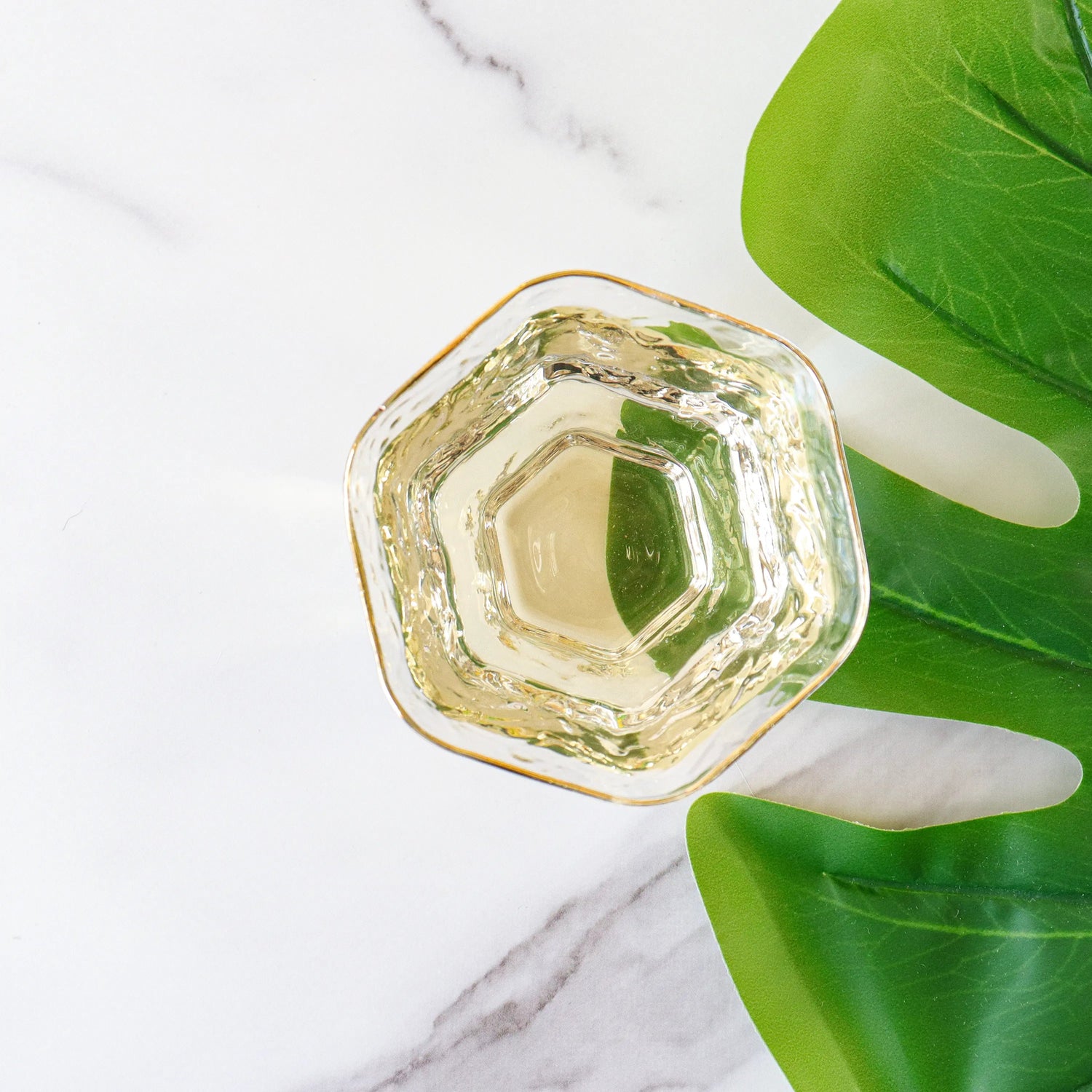 A hexagonal glass filled with Tie Guan Yin Oolong Tea by My Store, its clear liquid subtly evoking an orchid aroma, rests on a white marble surface beside a large green leaf.