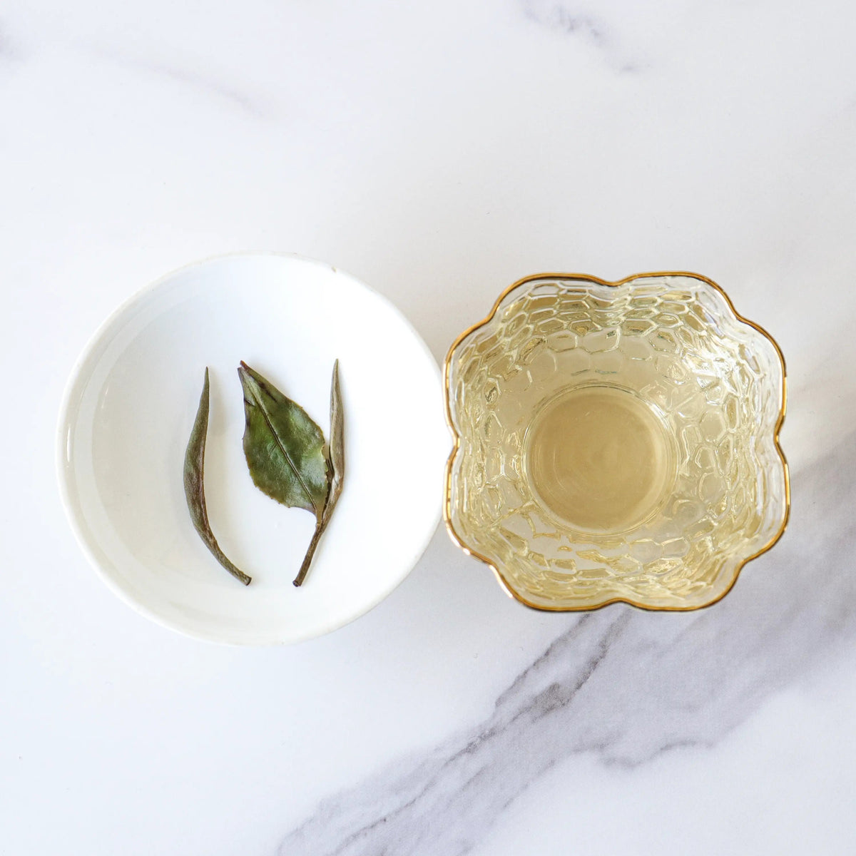White Peony tea leaves beside a gold-rimmed glass cup of pale tea on marble.