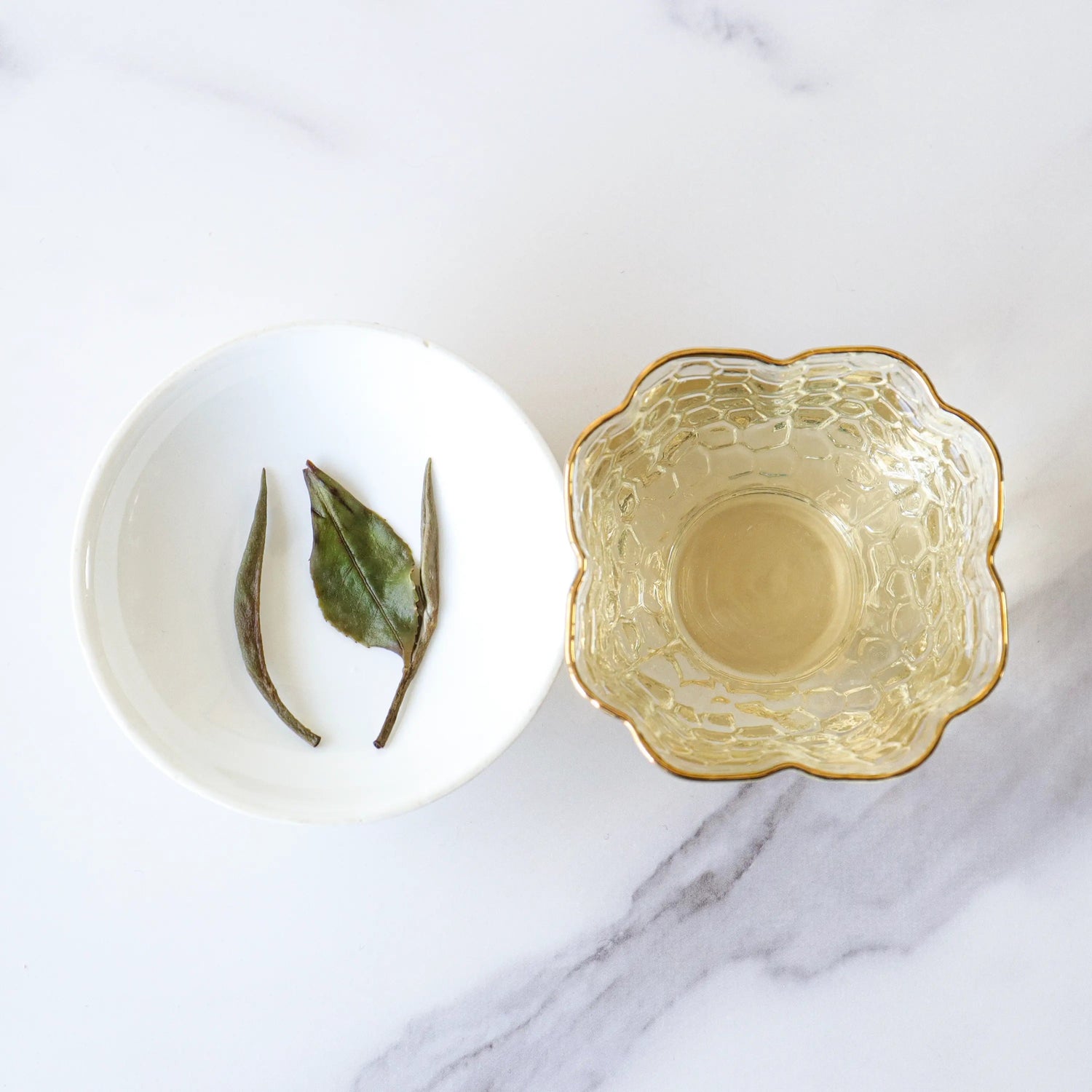 A white bowl with three dried My Store White Peony Tea leaves sits beside a clear, flower-shaped glass cup filled with pale White Peony Tea from My Store on a marble surface.