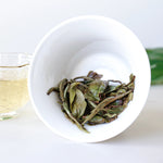 A white cup on its side holds wet White Peony Tea leaves from My Store, with a glass of pale white tea in the background.