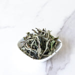 A small white dish filled with loose dried My Store White Peony Tea leaves sits on a white marble surface.
