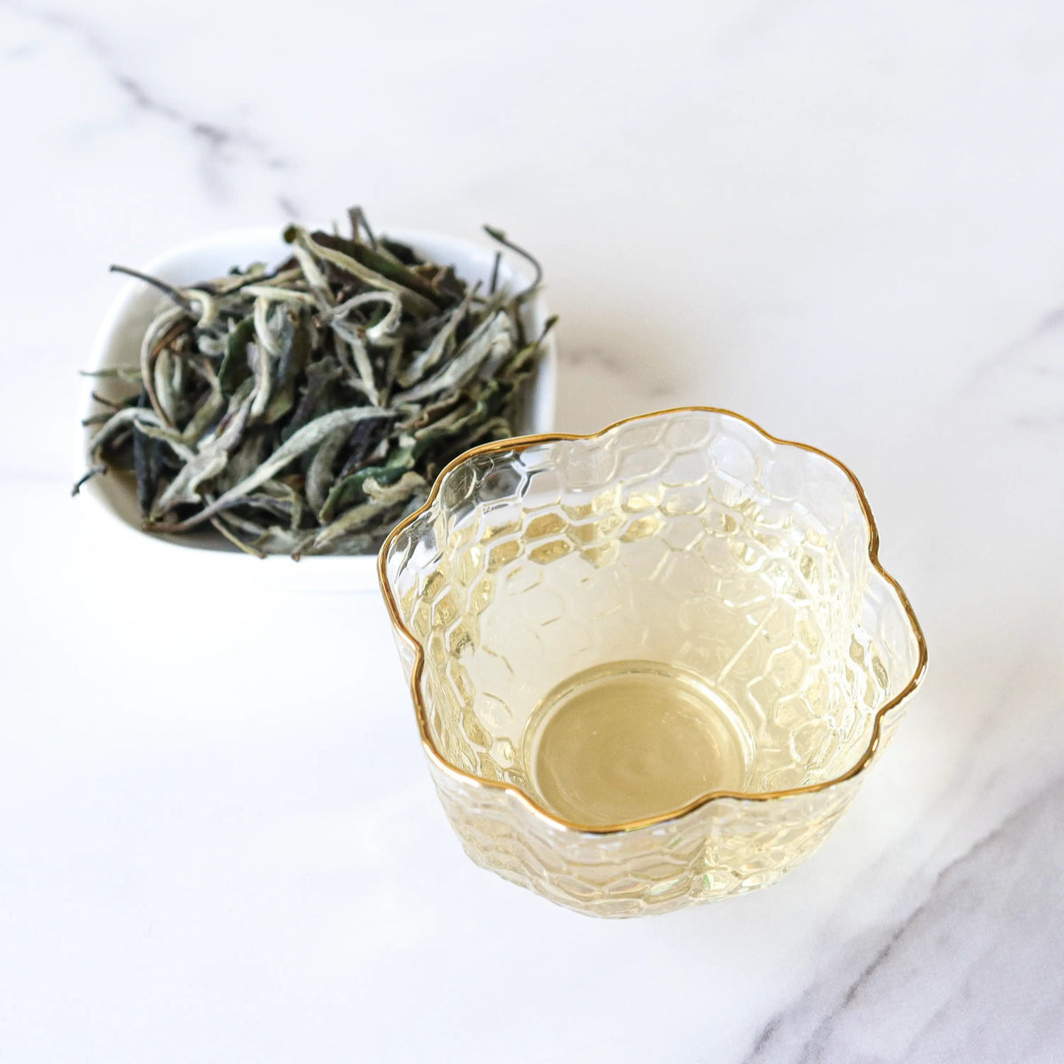 White Peony tea leaves beside a gold-rimmed glass cup of pale tea on marble.