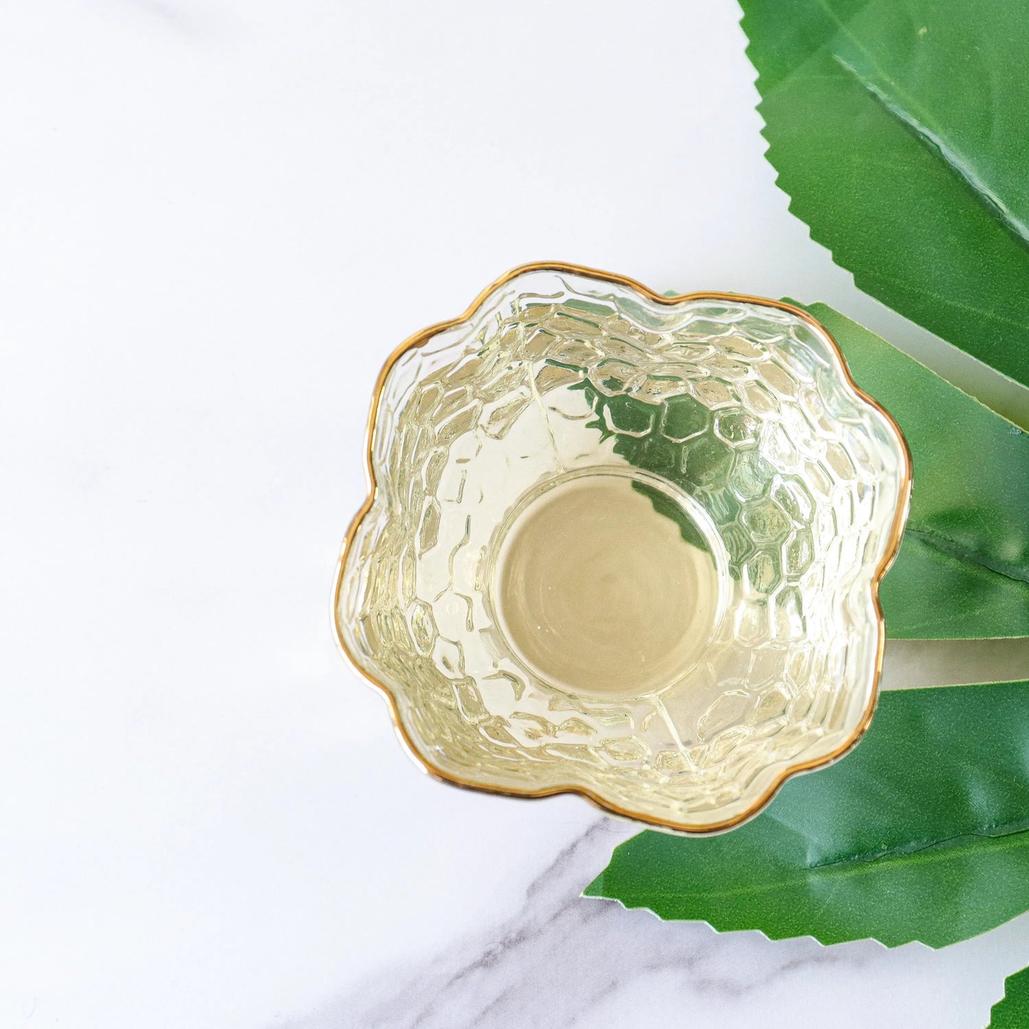 A gold-rimmed, textured glass bowl in a flower shape is photographed from above beside green artificial leaves and White Peony Tea by My Store on a white marble surface.