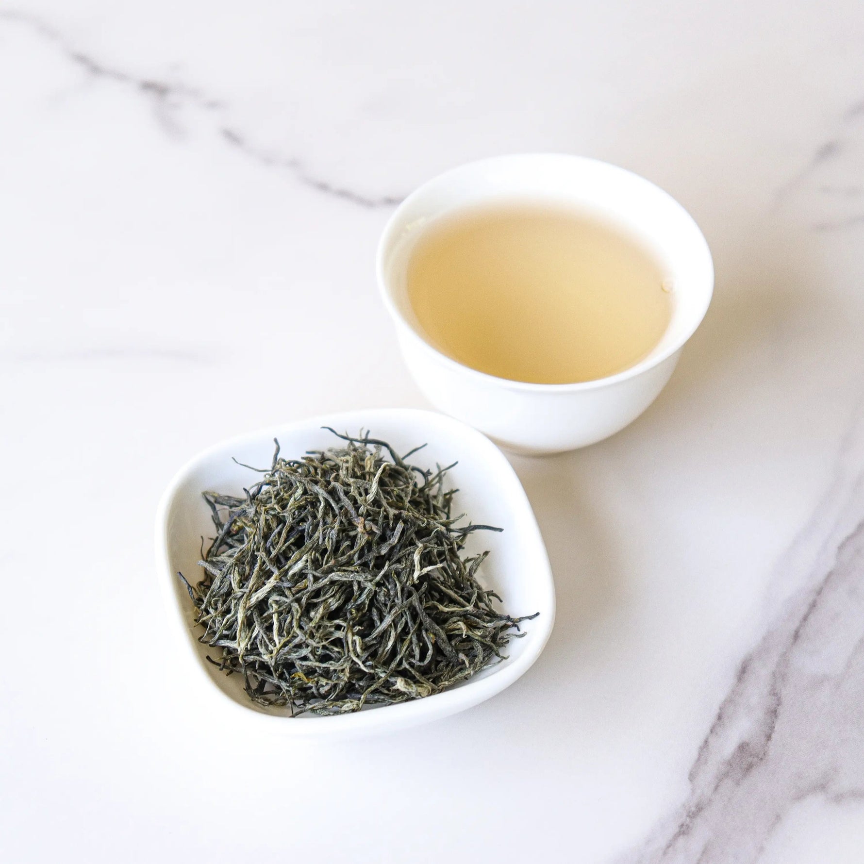 A small white bowl holds Xinyang Maojian Green Tea from My Store, placed next to a white cup of light yellow tea on a marble surface.
