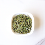 A white bowl filled with loose, dried Zhu Ye Qing Green Tea leaves from My Store sits on a white surface—a premium Chinese tea valued for its wellness benefits.