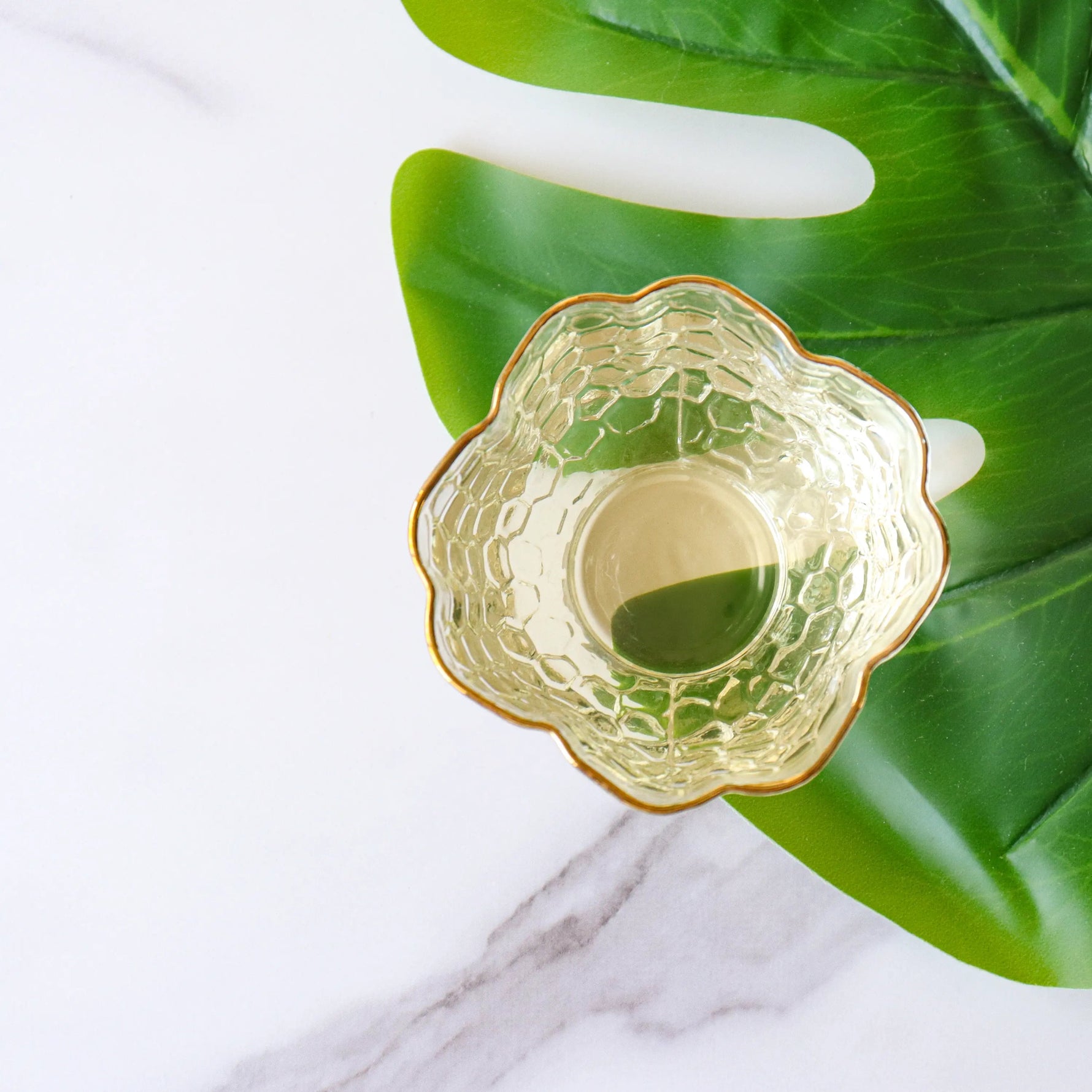 A textured glass bowl rests on a green leaf atop white marble, capturing the freshness of My Store's Zhu Ye Qing Green Tea, renowned for wellness benefits.